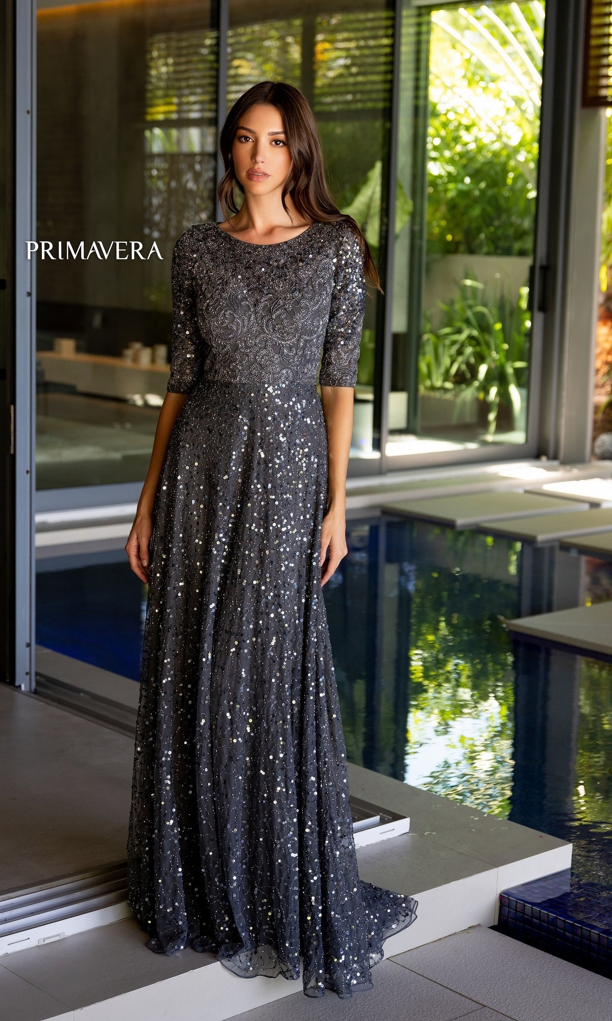 A woman stands indoors by a pool wearing the Long Prom Dress 13115 by Primavera, a floor-length silver-gray A-line beaded formal dress with half sleeves. The word